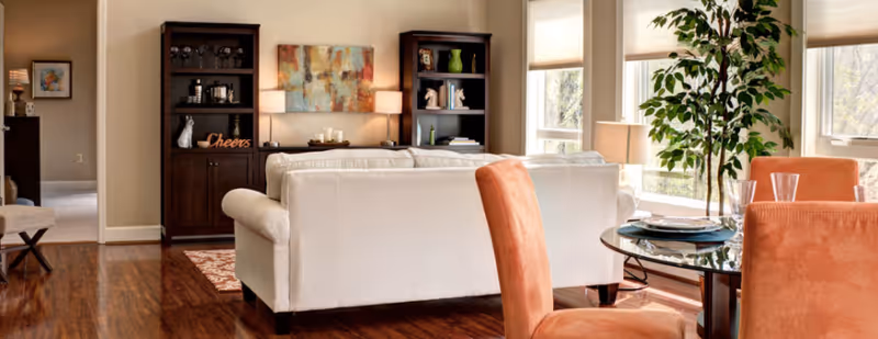 A bright living room with a white sofa facing two dark wooden bookshelves and a colorful abstract painting on the wall. To the right, there is a round glass dining table with orange upholstered chairs and a tall green plant near large windows letting in natural light. The floor is wooden and there is a small rug partially visible under the sofa.