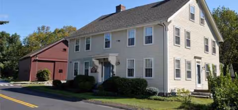 Exterior view of Carleton-Willard Village building, two-story beige siding with a red annex along a road.