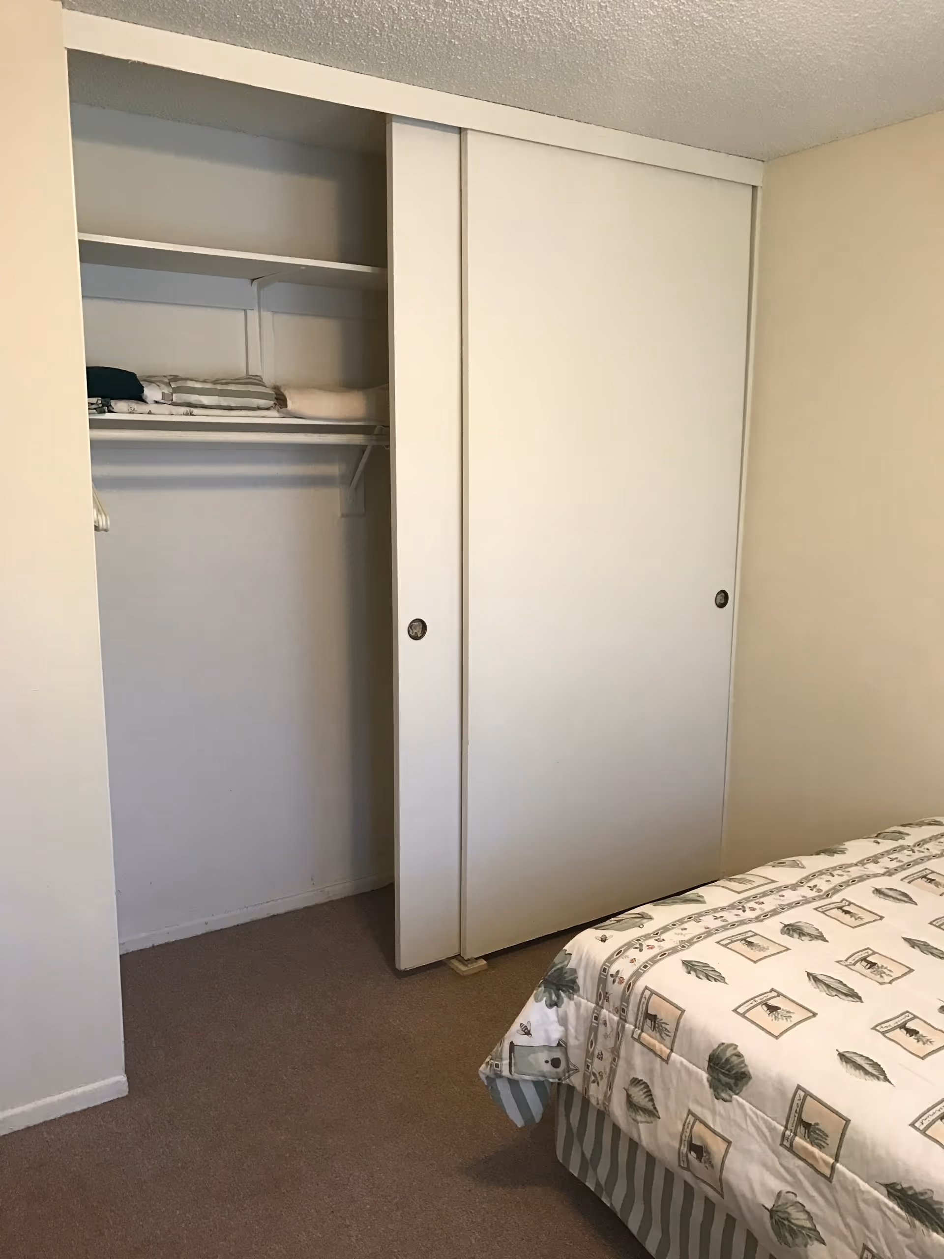 A bedroom corner showing a partially open white sliding closet door with shelves inside holding folded towels and clothes. Part of a bed with a patterned bedspread featuring leaves and framed images is visible on the right side. The walls are beige and the floor is carpeted.