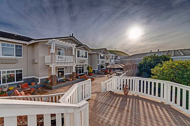 Sunlit outdoor wooden deck and seating courtyard in front of a two-story senior living building with balconies and landscaping.
