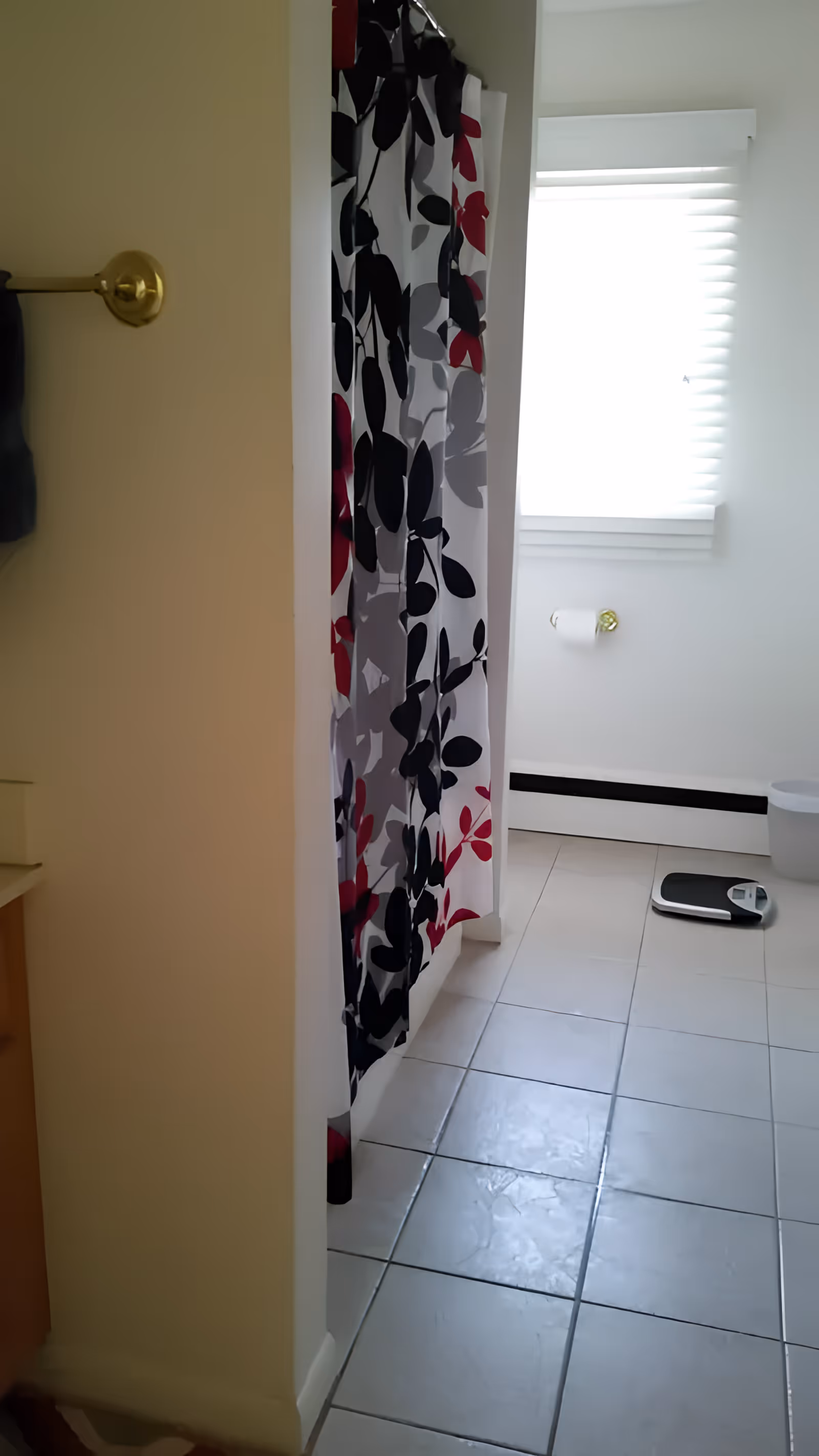 A bathroom with a tiled floor, a shower with a curtain featuring a black, gray, and red leaf pattern, a window with white blinds, a toilet paper holder with a roll of toilet paper, a white trash bin, and a digital scale on the floor.
