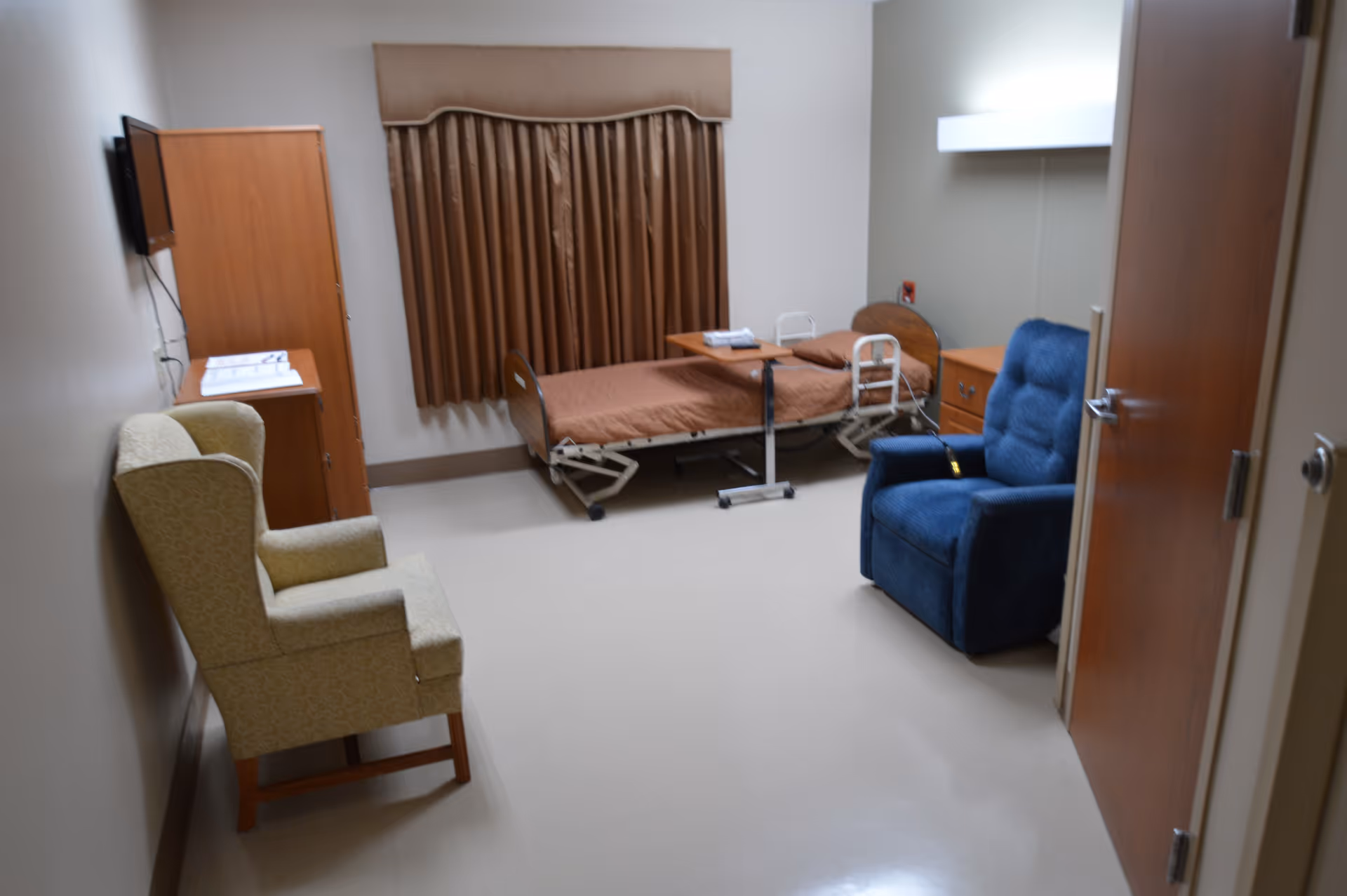 A small room with a hospital bed covered in a brown blanket, a blue recliner chair, a beige armchair, a wooden cabinet, a small table with papers, and a window with brown curtains.