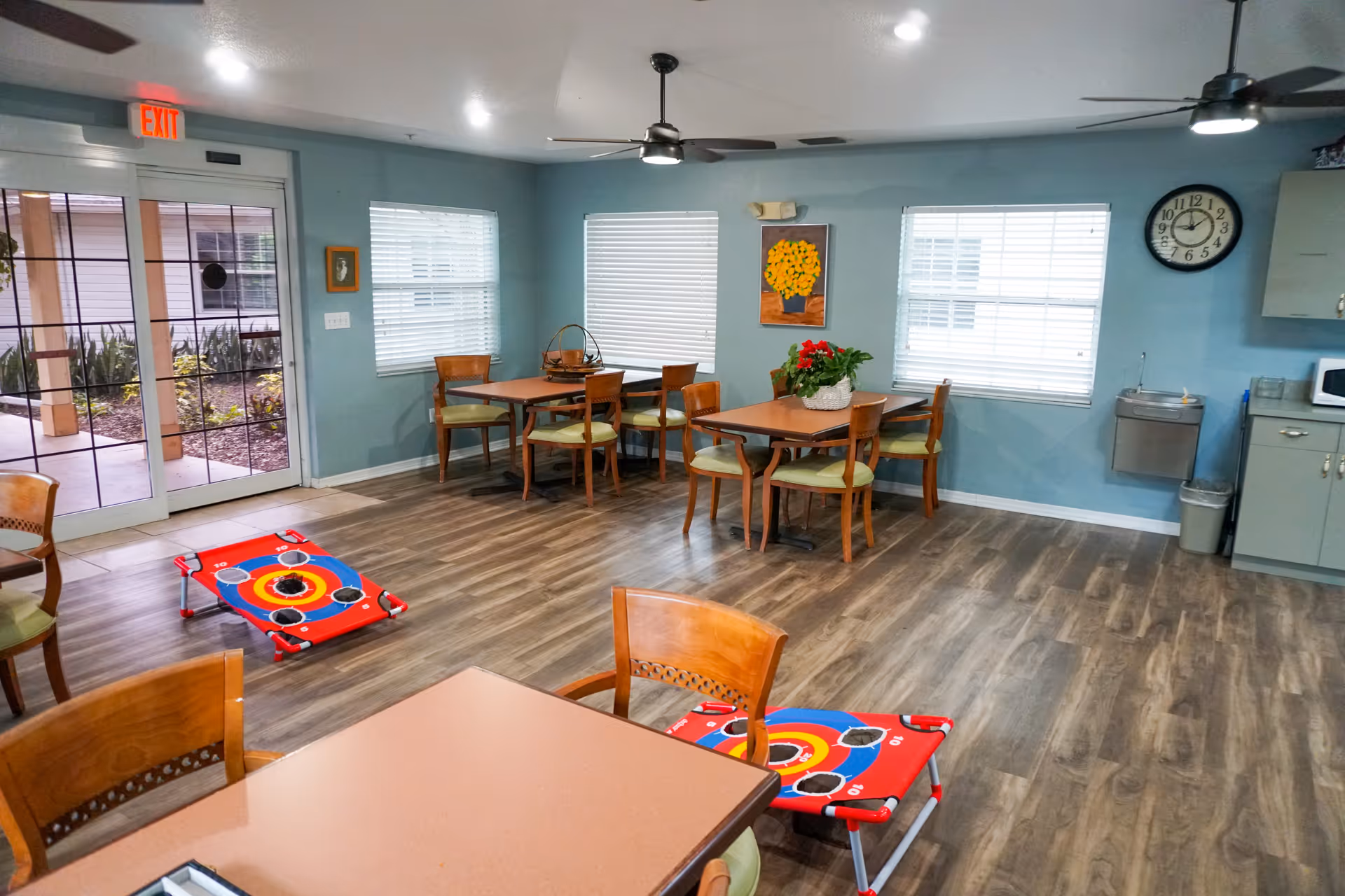 A bright common area with light blue walls and wood flooring featuring several wooden tables and chairs with green cushions. There are two red bean bag toss games on the floor. The room has three windows with white blinds, a wall clock, a water fountain, a microwave on a counter, ceiling fans, and an exit door leading outside to a garden area.