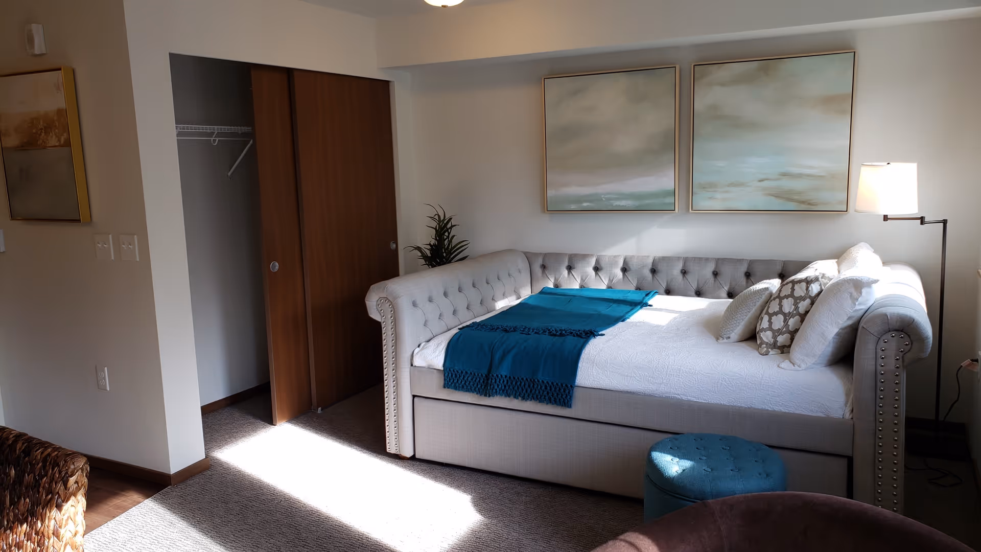 A cozy bedroom with a light gray tufted daybed adorned with white and patterned pillows and a teal throw blanket. Above the bed are two abstract paintings in soft, muted tones. To the right of the bed is a floor lamp with a white shade. On the left side, there is an open closet with wooden sliding doors and a small green plant beside the bed. The room has beige carpeting and soft natural light coming in.
