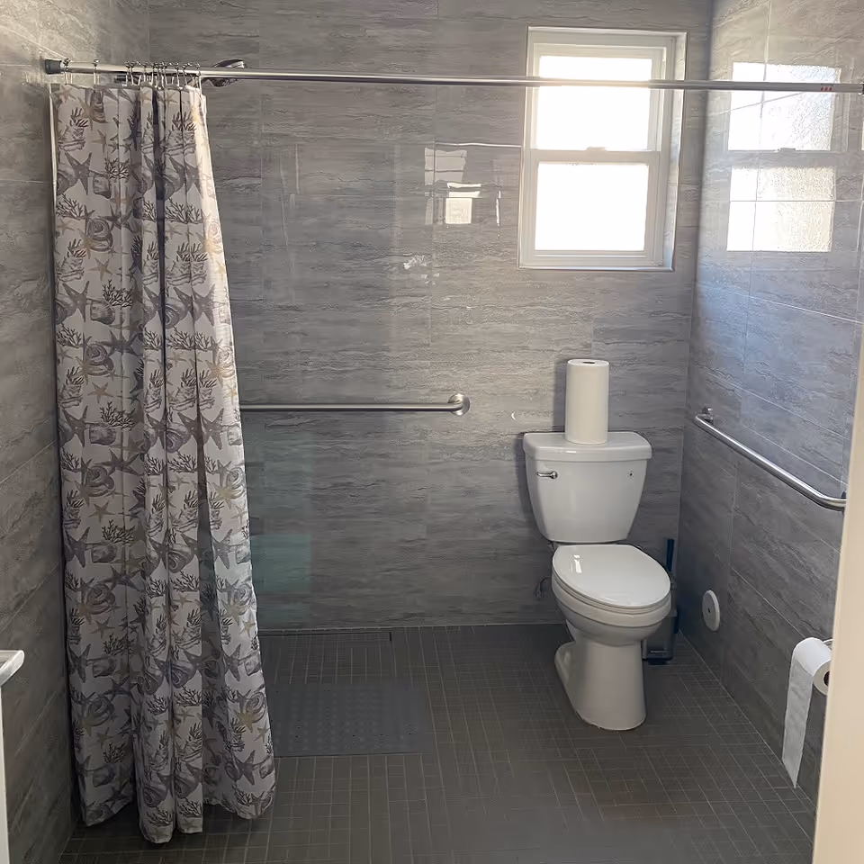 Accessible bathroom with a toilet, grab bars, a patterned shower curtain, and gray tiled walls and floor beneath a small window.