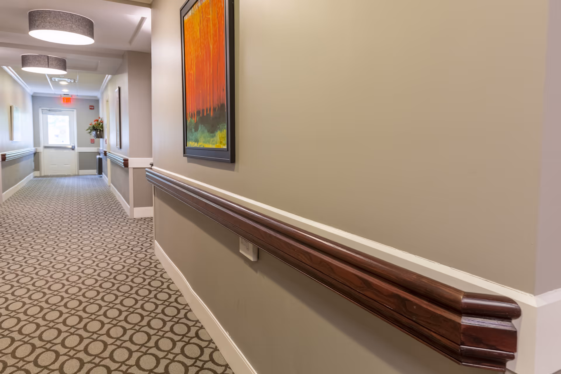 A well-lit hallway with patterned carpet, beige walls, wooden handrails on both sides, ceiling lights, and framed artwork on the walls. A door with a window is visible at the end of the hallway.