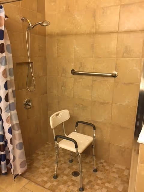 Shower area with beige tiled walls and floor, equipped with a handheld showerhead, a grab bar, and a white shower chair with armrests. A shower curtain with a polka dot pattern is partially visible on the left side.