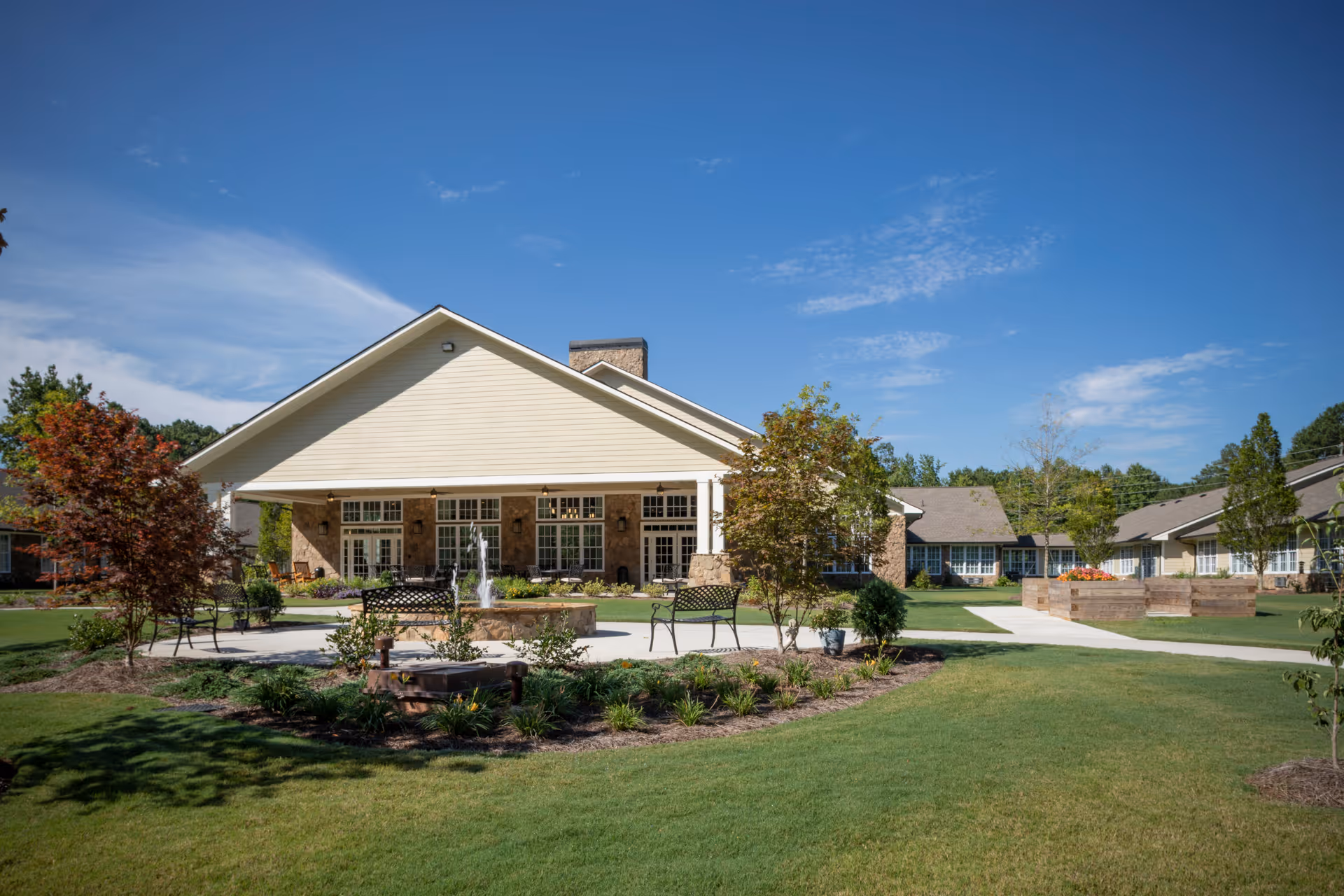 Front exterior of a single-story assisted living building with a central fountain, benches, and landscaped lawns under a blue sky.