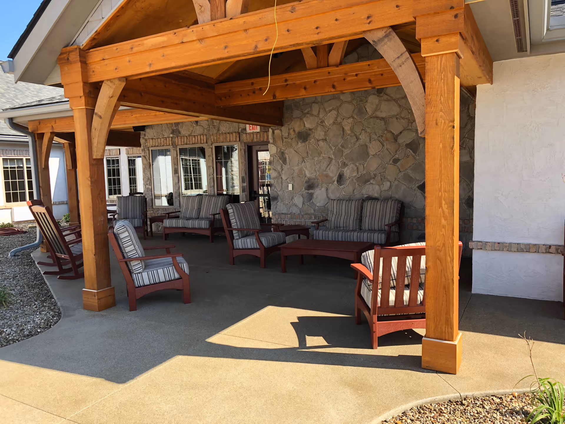 Covered outdoor patio with wooden beams, a stone wall, and striped cushioned chairs and benches.