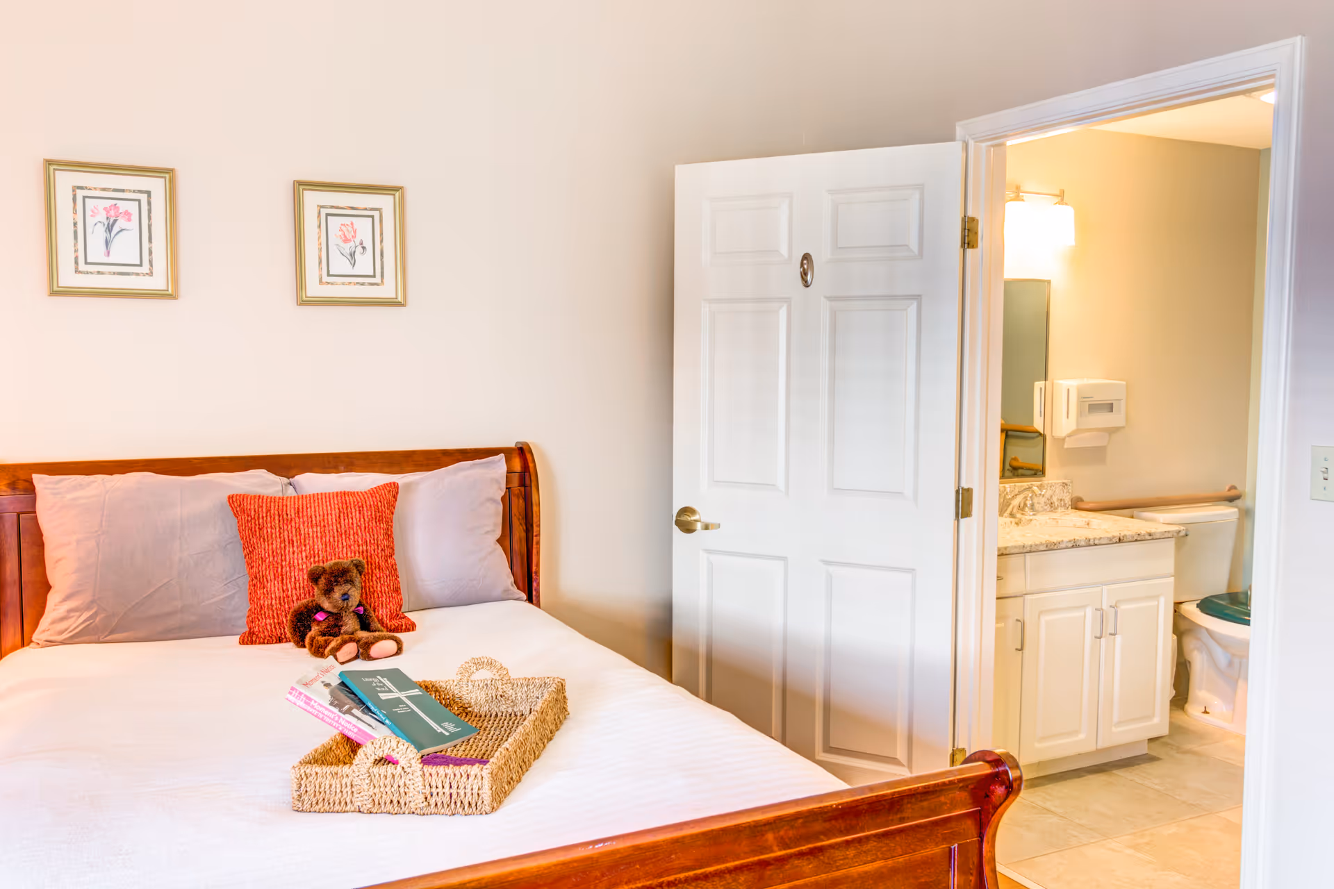 A cozy bedroom with a wooden bed frame, white bedding, two pillows, a red decorative pillow, and a small teddy bear. A woven tray with books is placed on the bed. The door to an adjacent bathroom is open, showing a vanity with a granite countertop, a mirror, and a toilet.