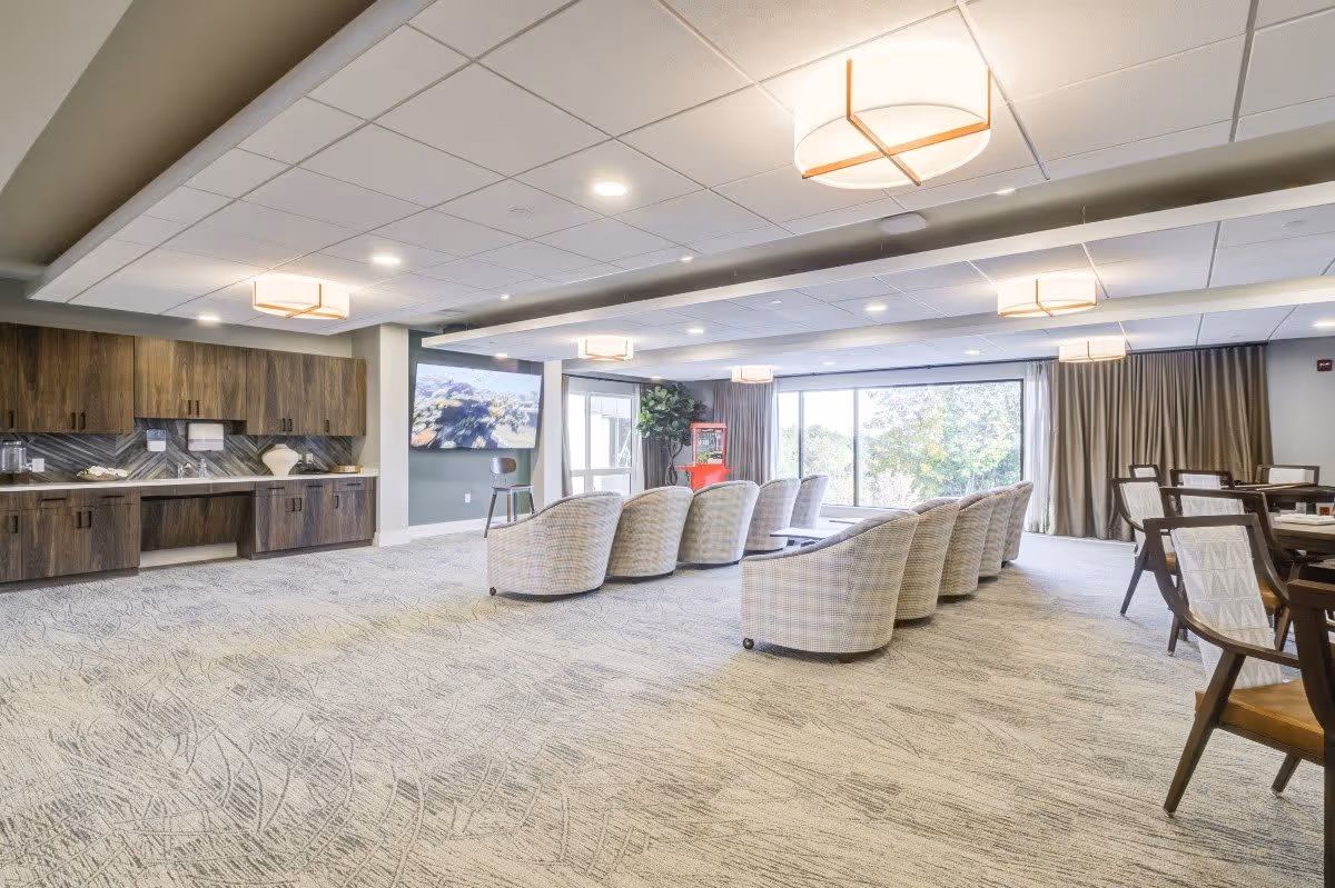 Spacious senior living common lounge with upholstered chairs arranged facing a TV, dining tables, built-in cabinets, large windows, and ceiling lights.