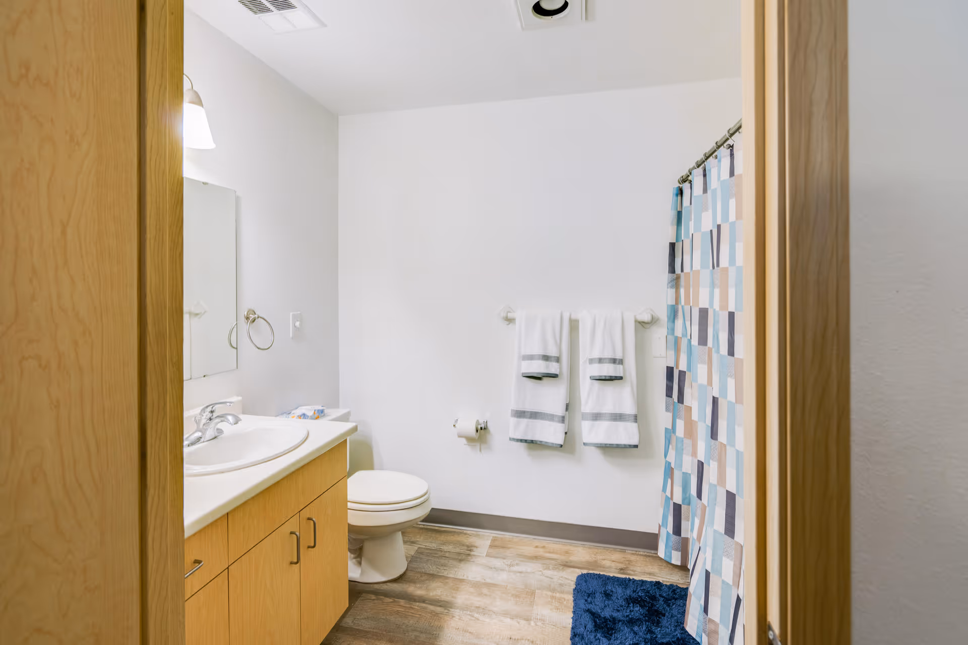 A clean bathroom with a wooden vanity and white sink on the left, a toilet in the center, two white towels hanging on a towel rack on the wall, and a shower with a colorful checkered shower curtain on the right. The floor has wood-like tiles and there is a blue bath mat in front of the shower.