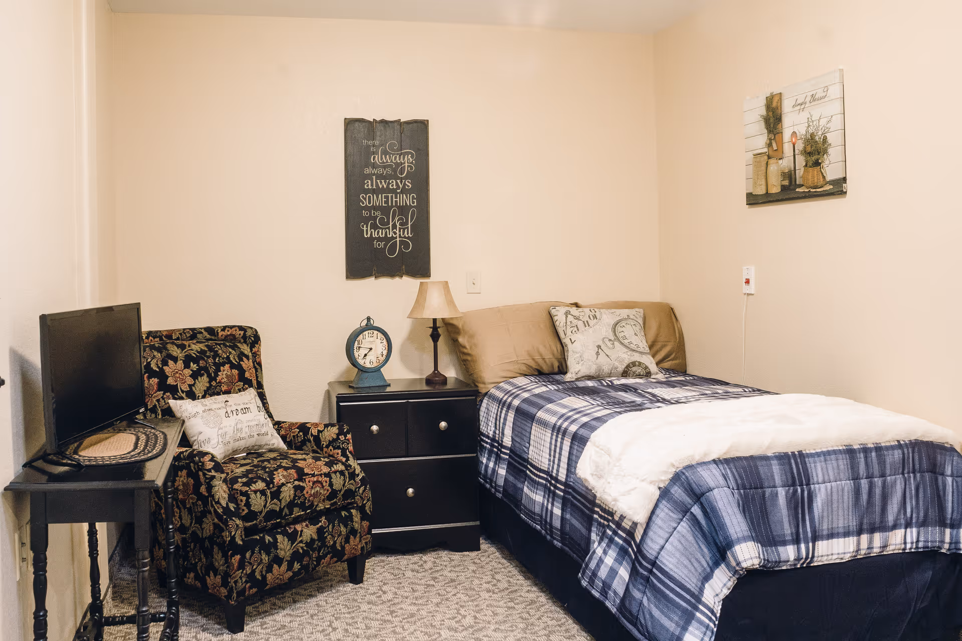 A cozy bedroom with a single bed covered in a blue plaid blanket and beige pillows. Next to the bed is a black nightstand with a vintage-style clock and a small lamp. A floral upholstered armchair with a decorative pillow sits beside a small black table holding a flat-screen TV. The walls are decorated with two framed artworks, one with an inspirational quote and the other with a rustic design.
