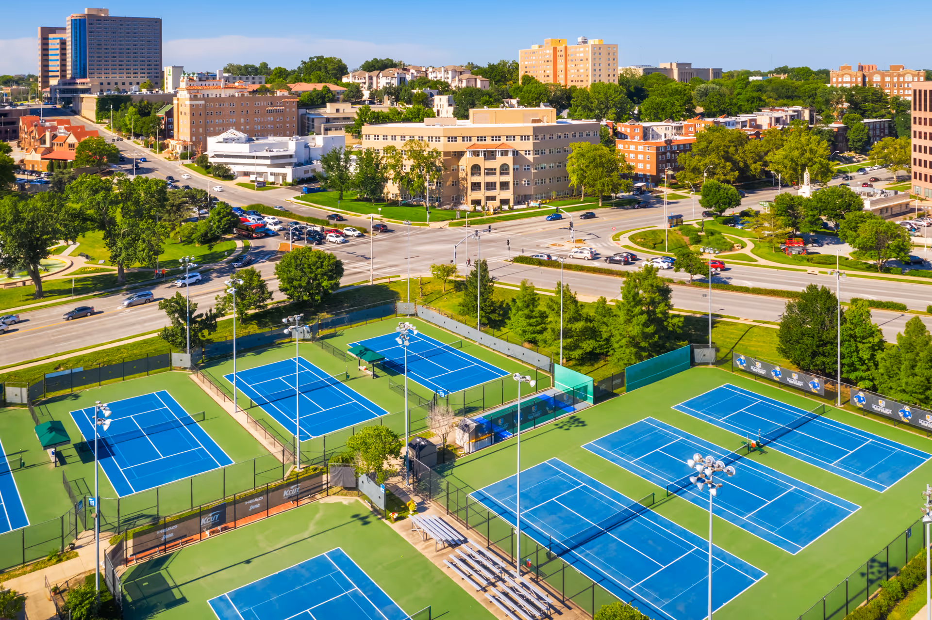 Aerial view of multiple blue tennis courts surrounded by green fencing and trees, with a cityscape of buildings, roads, and parked cars in the background under a clear sky.