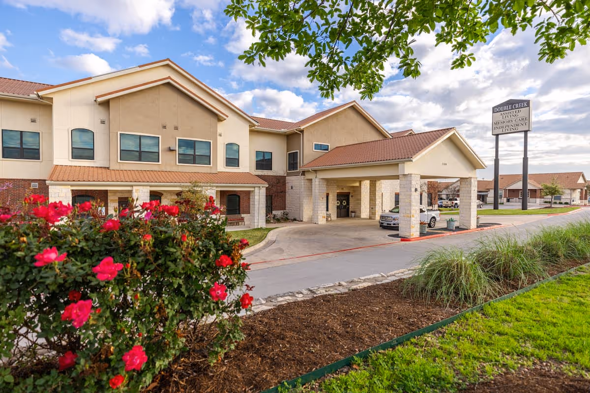 Exterior view of Double Creek Assisted Living and Memory Care facility showing a two-story building with a covered entrance driveway, a sign with the facility name, and landscaping including red flowers and green grass under a partly cloudy sky.