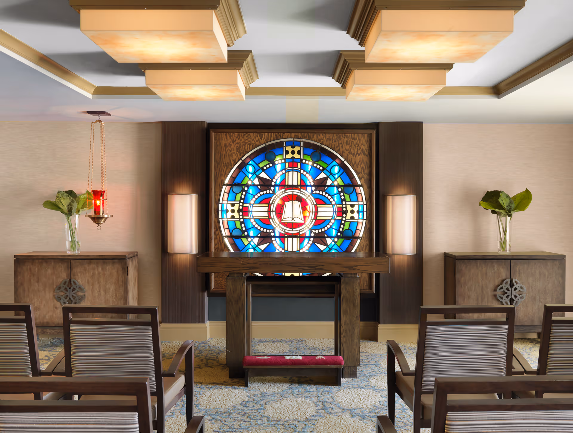 Interior view of a small chapel or meditation room with wooden chairs arranged in rows facing a wooden altar. Behind the altar is a large, colorful stained glass window with a circular design featuring a book at the center. Two wooden cabinets with vases holding green plants flank the altar. The ceiling has decorative light fixtures.