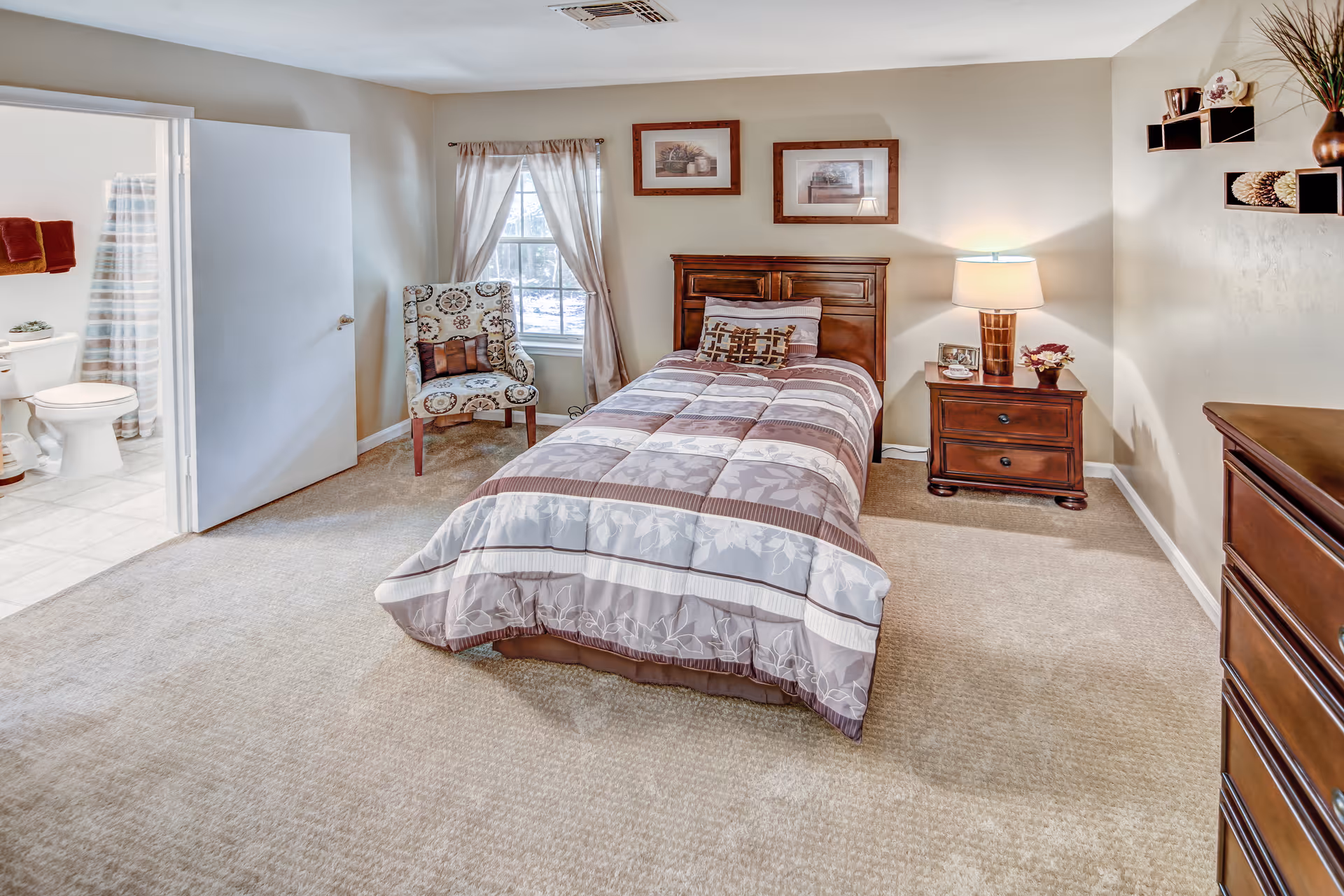 A bright furnished bedroom with a single bed, nightstand, dresser, armchair, and an open door revealing a bathroom.