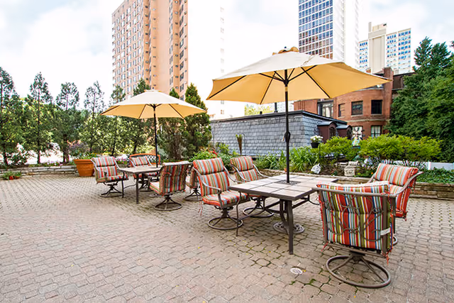 Outdoor patio area with several tables and chairs featuring striped cushions, each table shaded by a large beige umbrella. The patio is paved with bricks and surrounded by greenery and tall buildings in the background.