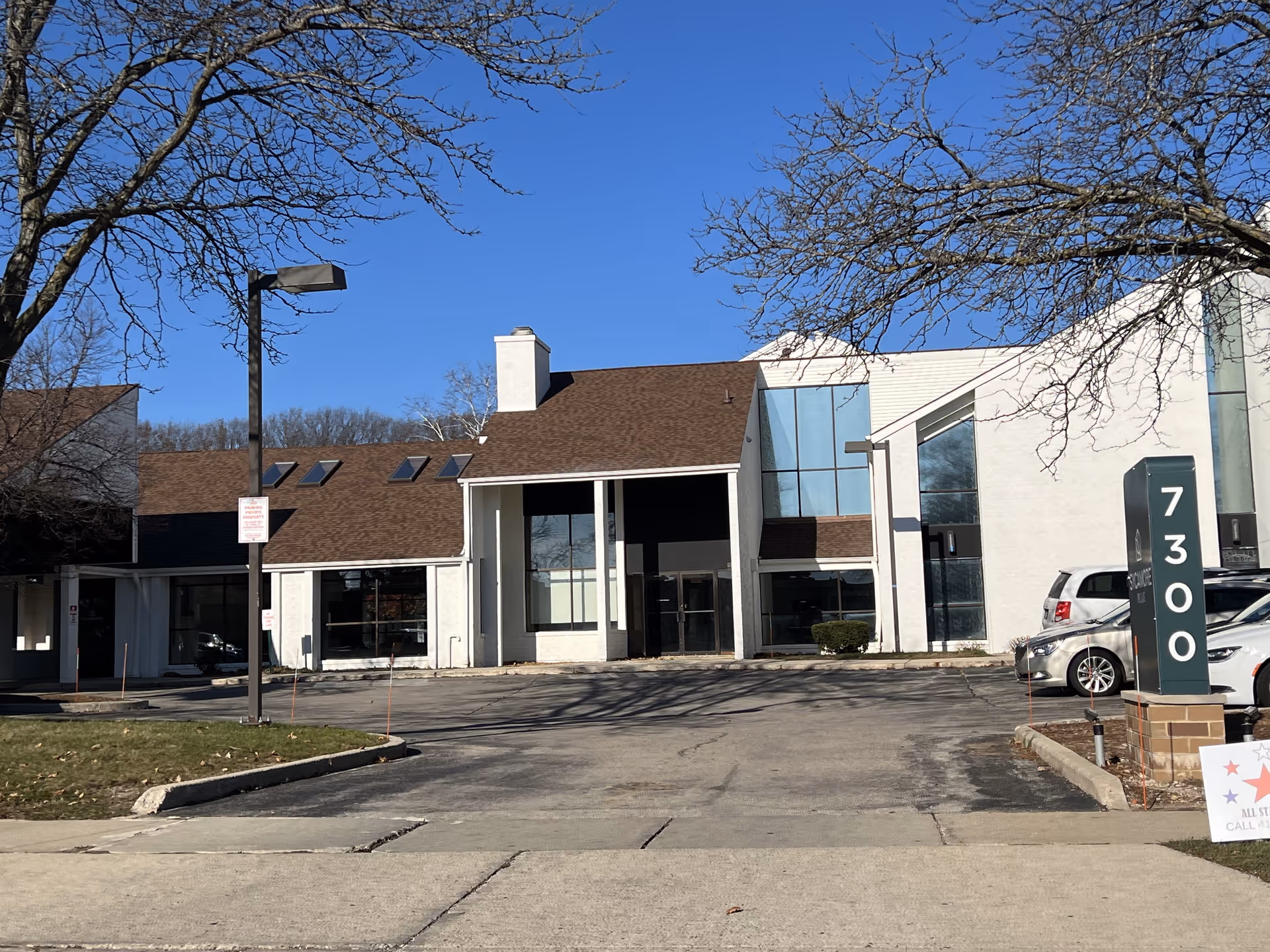 Exterior view of a senior living facility building with white walls and brown roof under a clear blue sky. There are leafless trees on both sides, a parking lot in front with several parked cars, and a green signpost displaying the number 7300.