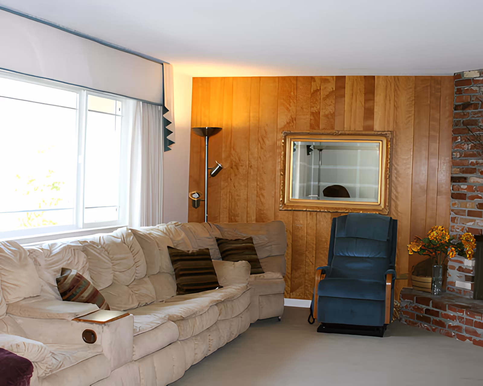 A cozy living room with a large beige sectional sofa adorned with striped pillows, a blue recliner chair, a floor lamp, a wooden paneled wall with a framed mirror, and a brick fireplace with a vase of yellow flowers.