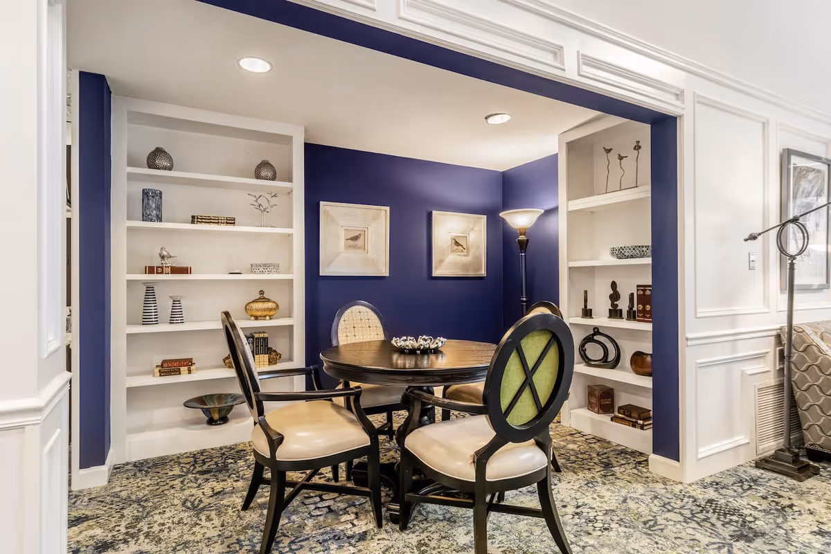 A cozy interior seating area with a round wooden table surrounded by four cushioned chairs. The walls are painted deep blue with white built-in shelves on either side filled with decorative items and books. Two framed bird pictures hang on the back wall, and a floor lamp provides soft lighting. The floor is covered with a patterned carpet.