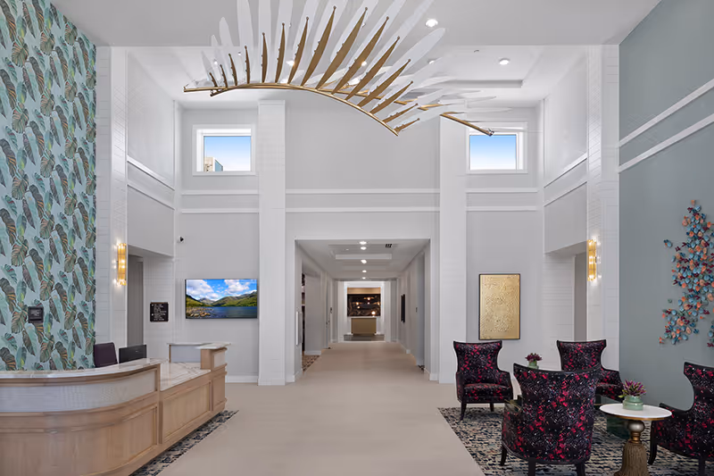 Bright modern lobby with a reception desk, high ceilings, sculptural chandelier, seating area with patterned chairs, and decorative wall art.