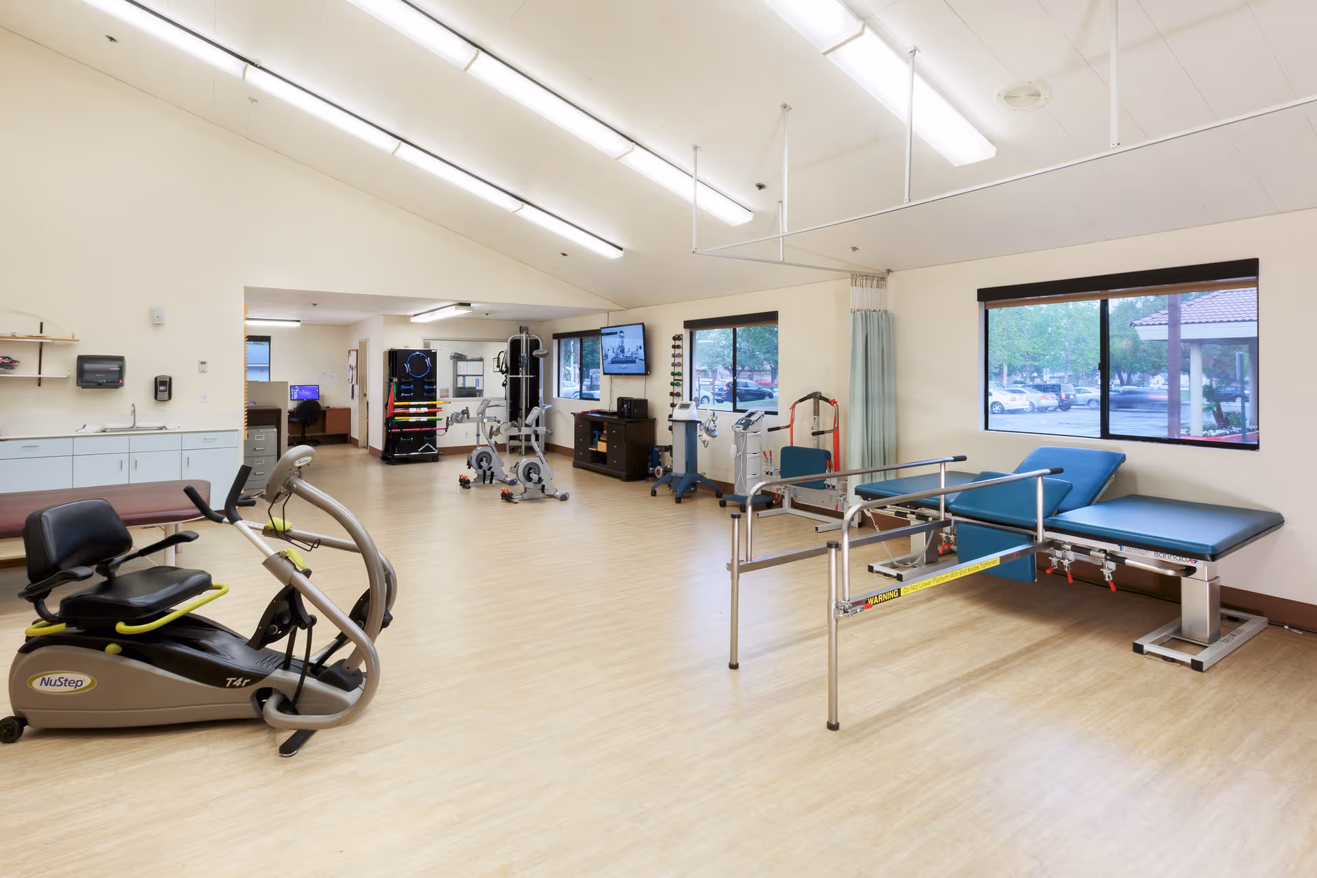 A spacious rehabilitation room with exercise equipment including a NuStep recumbent cross trainer, stationary bikes, parallel bars for walking support, and a padded examination table. The room has large windows letting in natural light, light-colored wooden flooring, and white walls with overhead fluorescent lighting.