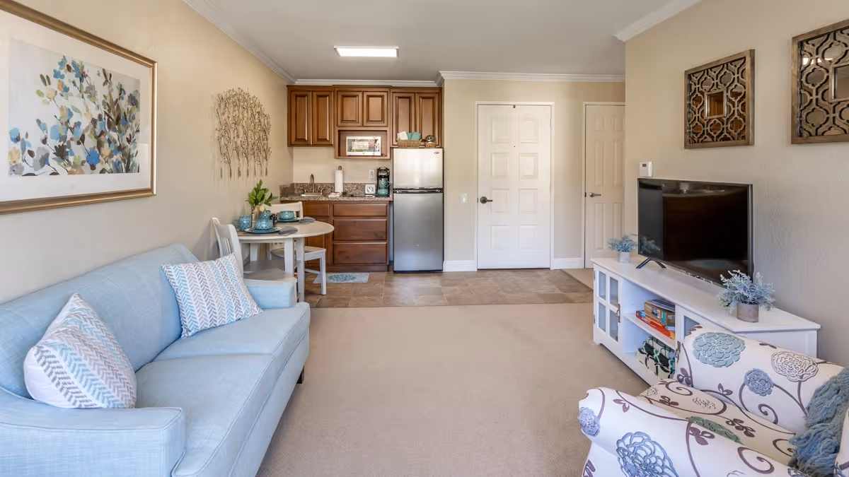A cozy living area in a senior living facility featuring a light blue sofa with patterned pillows, a small round dining table with two chairs, a kitchenette with wooden cabinets and a stainless steel refrigerator, and a white TV stand with a flat-screen television. The room is decorated with framed artwork, wall decor, and small plants, creating a warm and inviting atmosphere.
