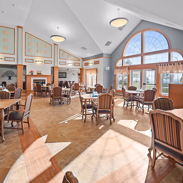 Spacious sunlit dining room with multiple tables and upholstered chairs, a high vaulted ceiling, large arched windows, and a fireplace.