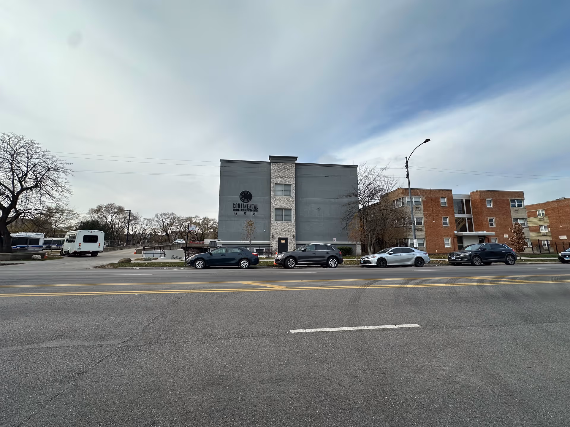 Exterior view of Continental Nursing & Rehabilitation Center, a multi-story building with a gray and brick facade. Several cars are parked along the street in front of the building, and there are leafless trees and a cloudy sky in the background.