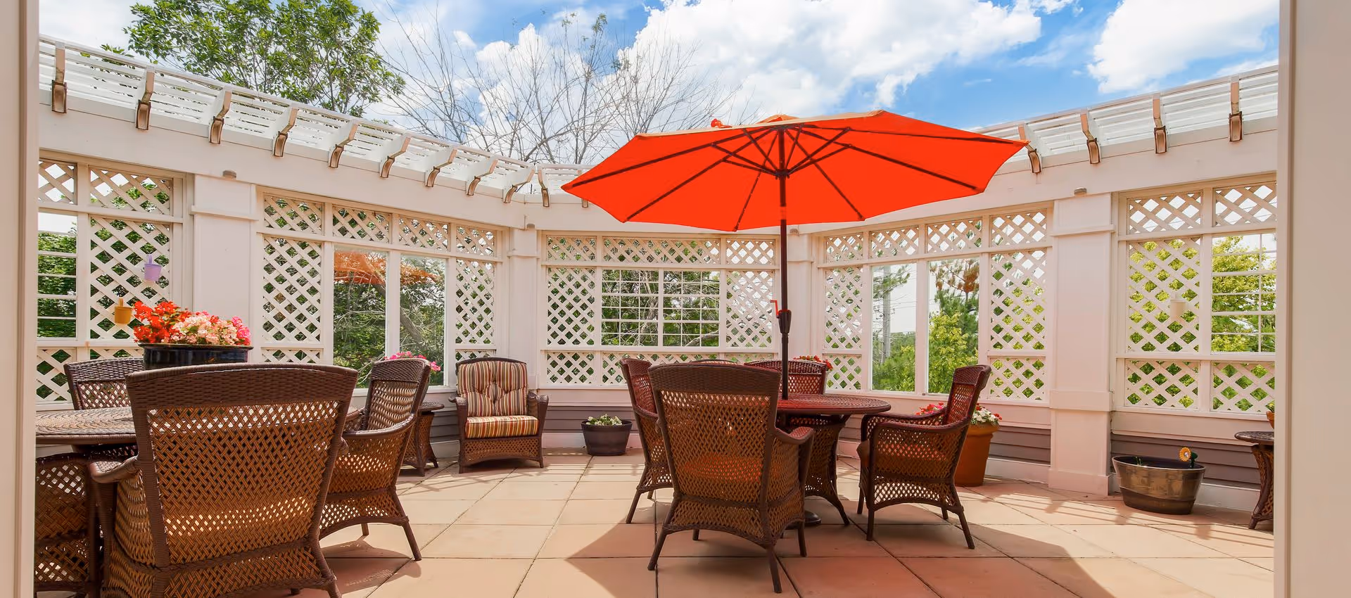 Outdoor patio area with wicker chairs and tables, one table featuring a large red umbrella. The patio is surrounded by white lattice fencing with some potted plants and flowers, under a partly cloudy blue sky.
