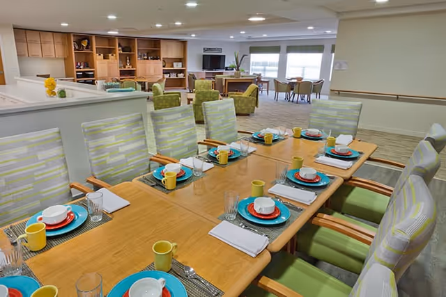 A well-lit dining area with a long wooden table set for a meal. The table has colorful plates, bowls, yellow mugs, glasses, and white napkins neatly arranged. Surrounding the table are cushioned chairs with light green and white striped upholstery. In the background, there is a lounge area with green armchairs, a TV, and large windows letting in natural light.