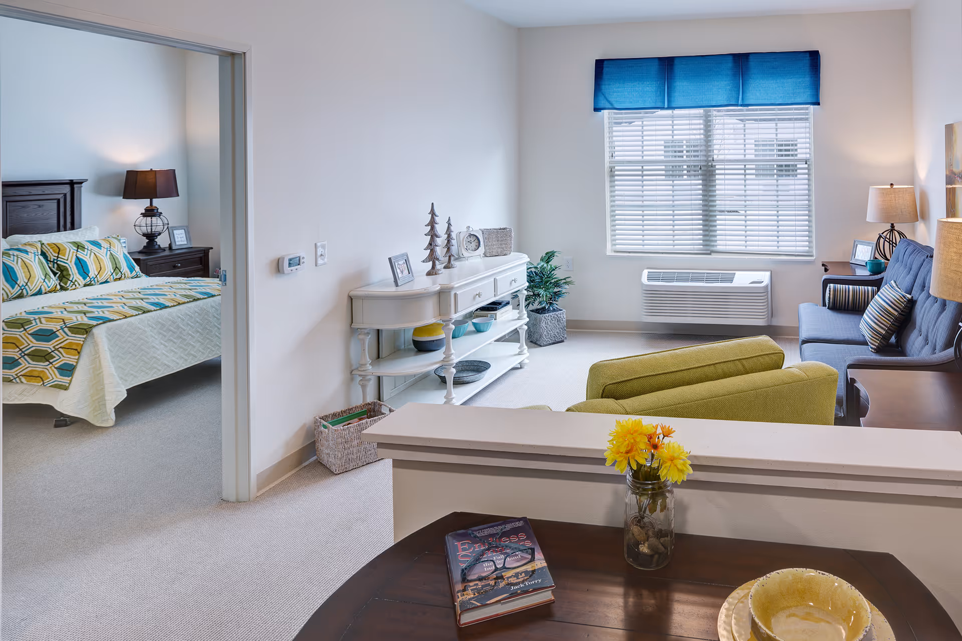 Bright furnished living room with green armchairs, a blue sofa, console table, and a view into a bedroom with patterned bedding.