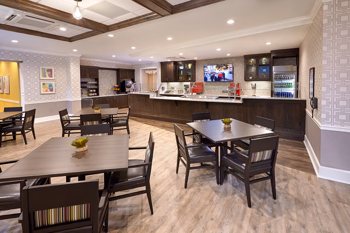 Bright communal dining area with multiple tables and chairs facing a long wood service counter and wall-mounted TV.