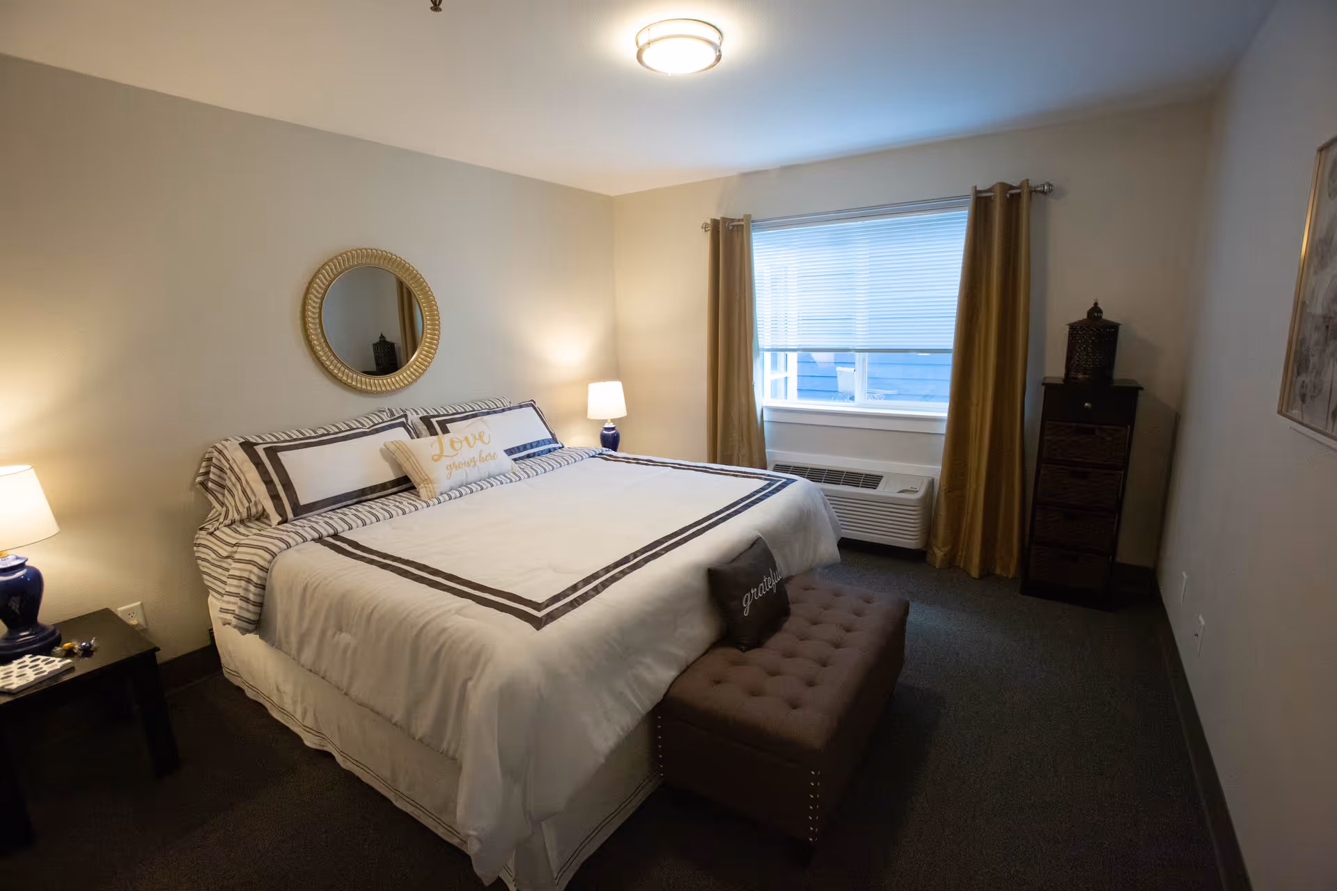 A cozy bedroom in Heatherwood Senior Living featuring a large bed with white and brown bedding, two pillows with inspirational words, a round mirror above the bed, two bedside tables with lamps, a window with beige curtains, a cushioned bench at the foot of the bed, and a tall chest of drawers in the corner.