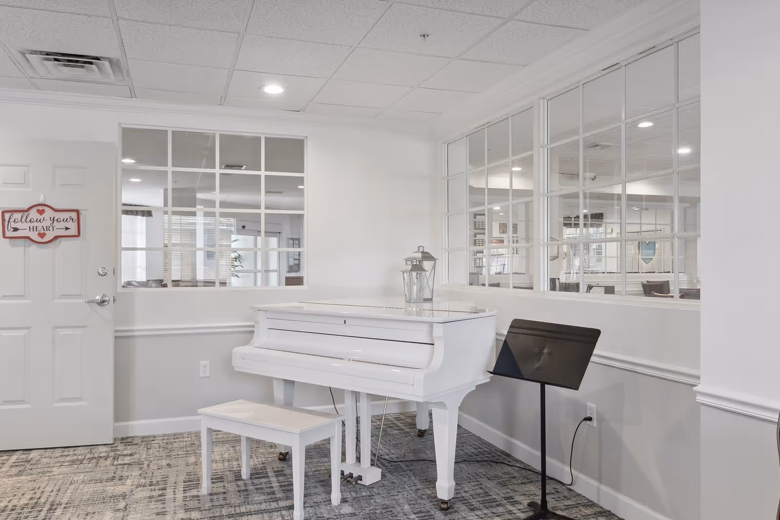 A bright room with a white grand piano and matching bench on a patterned carpet. The walls are white with large windows featuring multiple panes, allowing a view into an adjacent room. A music stand is positioned next to the piano. A door on the left has a decorative sign that reads 'follow your HEART'.
