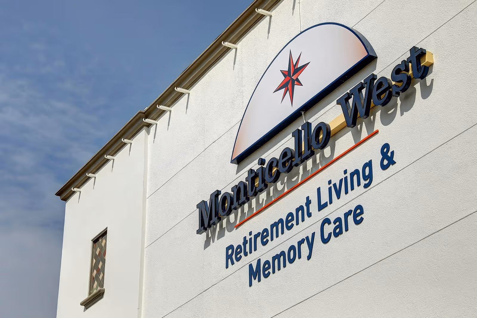 Facade of the Monticello West building showing the sign 'Monticello West Retirement Living & Memory Care' and a compass logo against a blue sky.