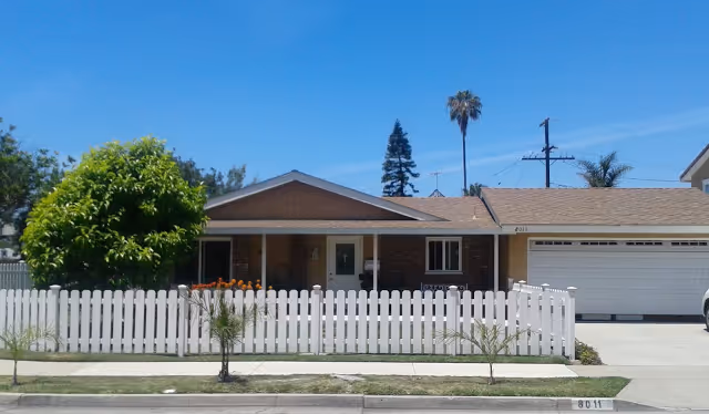 Single-story house with a white picket fence in front, a driveway leading to a two-car garage, and a small garden with a tree and flowers. The house has a brown roof and brick facade with a covered porch area.