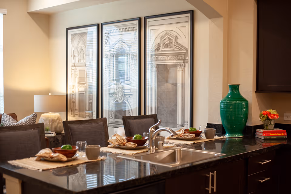 Kitchen island with a sink and place settings, a large green vase on the counter and a seating area with framed artwork in the background.