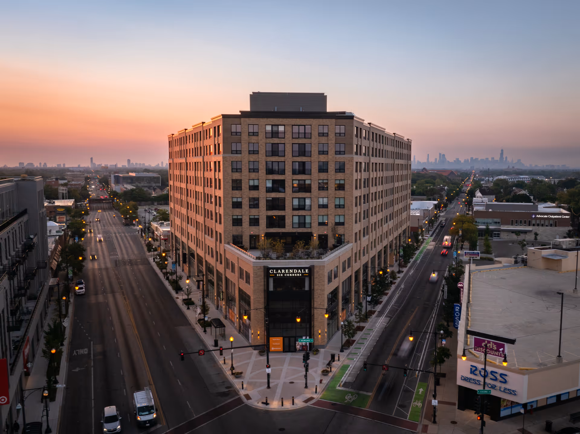Aerial view of a large multi-story brick building named Clarendale Six Corners at the intersection of two streets during sunset, with city skyline visible in the distance and streetlights illuminating the roads.