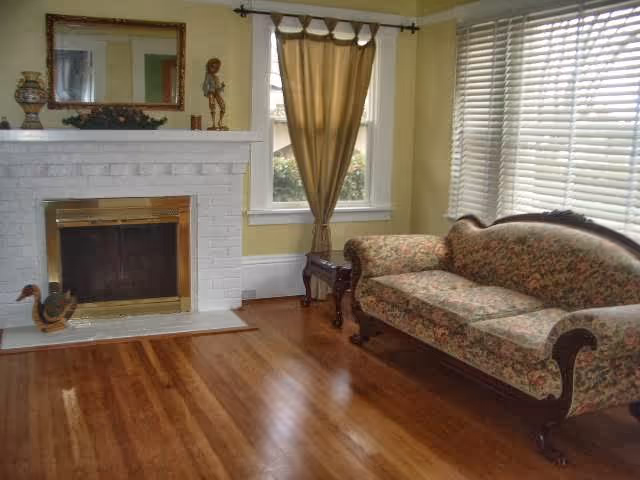 Sunlit living room with hardwood floors, a white brick fireplace, a floral upholstered sofa, and large windows with blinds and a curtain.