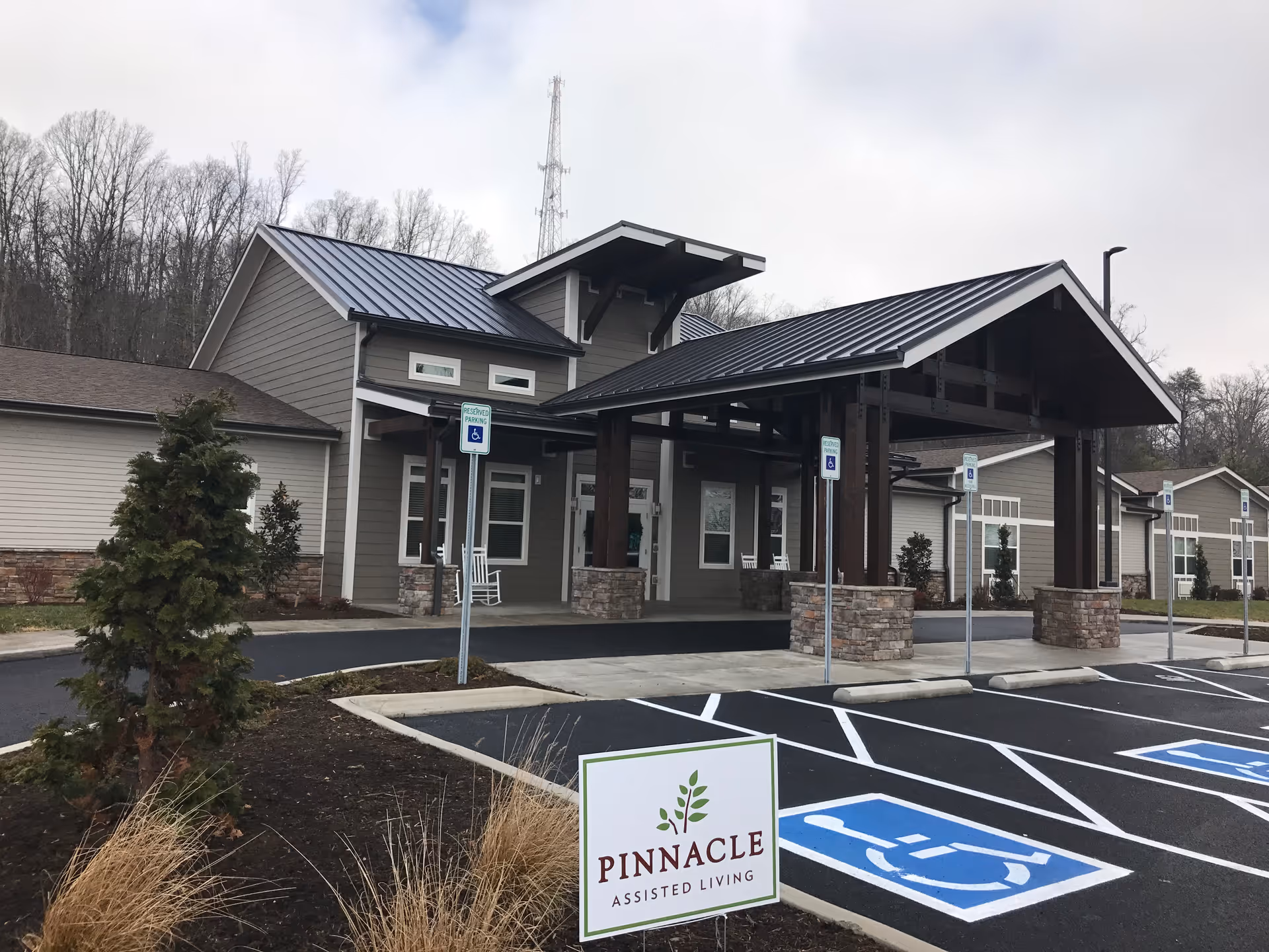 Exterior view of Pinnacle Assisted Living facility showing the entrance with a covered drop-off area supported by stone pillars, several handicap parking spaces in front, and a sign with the facility name. The building has a modern design with gray siding and a metal roof, surrounded by some landscaping and trees in the background.