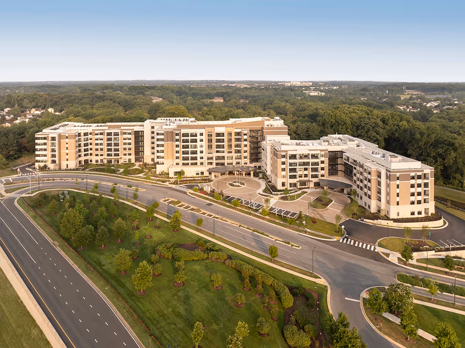 Aerial view of The Carnegie at Washingtonian Center, a large multi-story senior living facility surrounded by landscaped greenery and trees, with a curved driveway and adjacent roads.