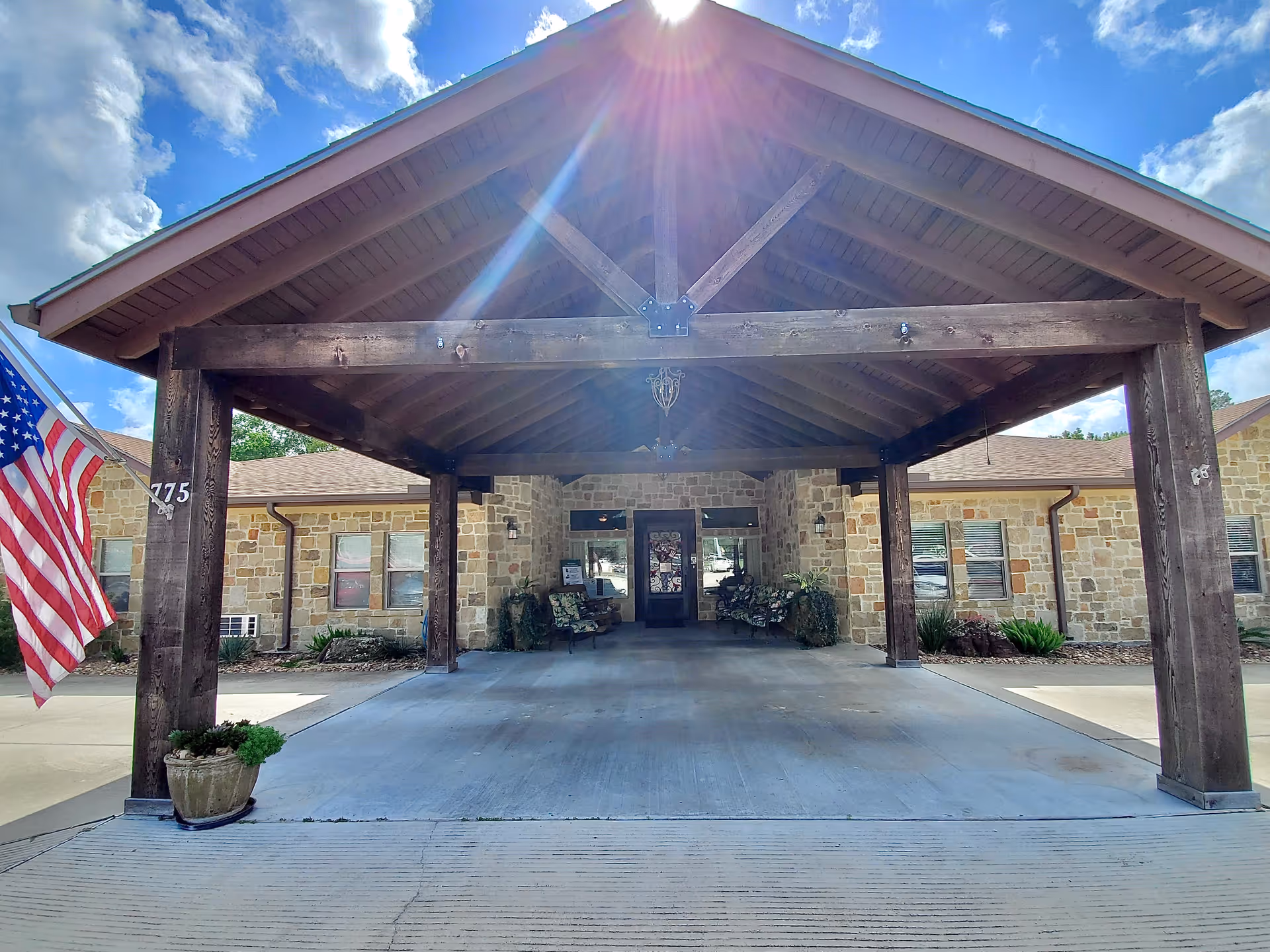 Covered entrance area of a building with wooden beams and a stone facade. There are benches with floral cushions on either side of the entrance door, potted plants, and an American flag on the left side. The sun is shining brightly above the roof.