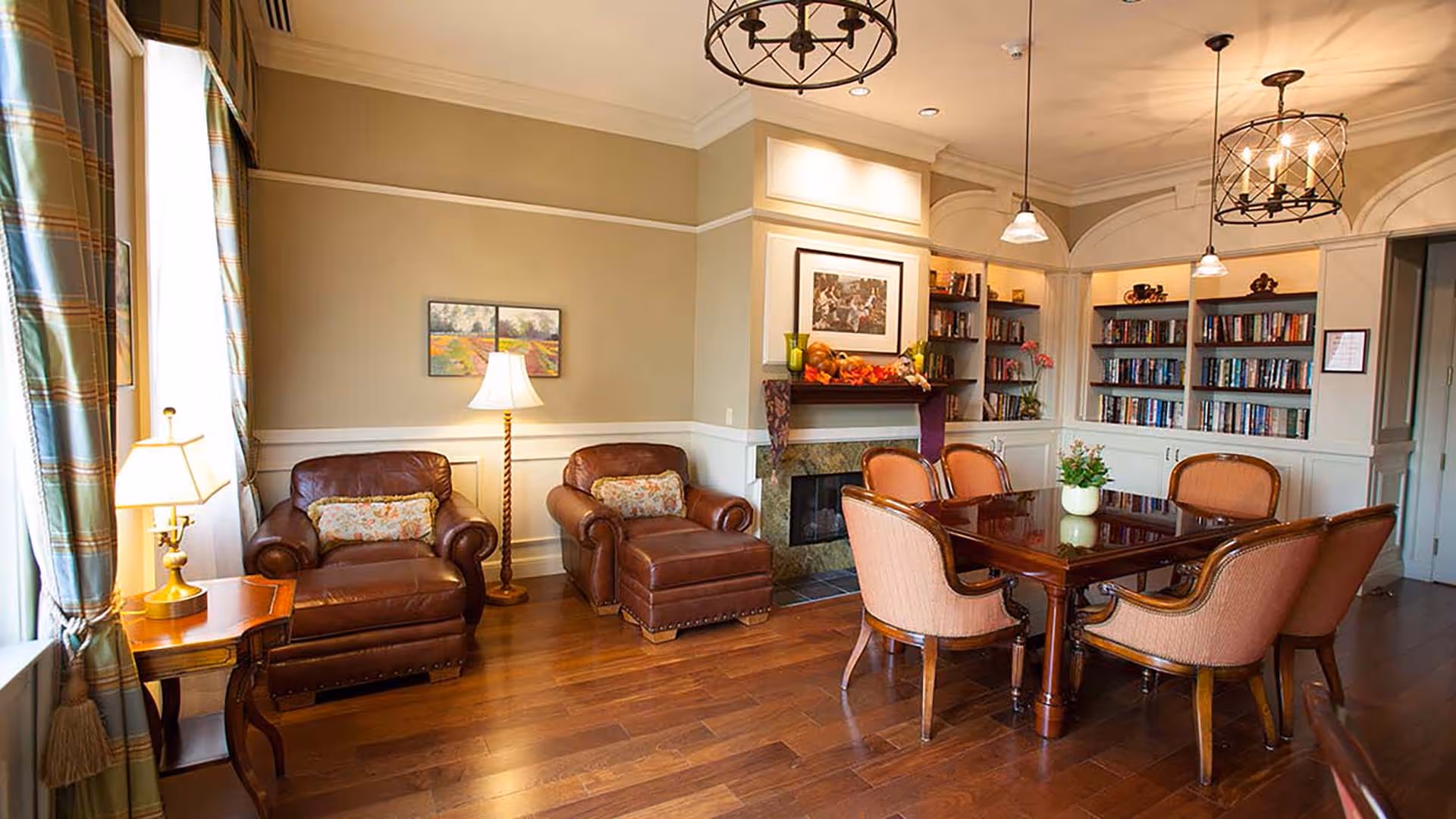 A cozy living room area with two brown leather armchairs and a matching ottoman, a wooden side table with a lamp, and a wooden dining table surrounded by six upholstered chairs. The room features hardwood floors, a fireplace with a mantel decorated with autumn-themed items, built-in bookshelves filled with books, and large windows with plaid curtains allowing natural light to enter.