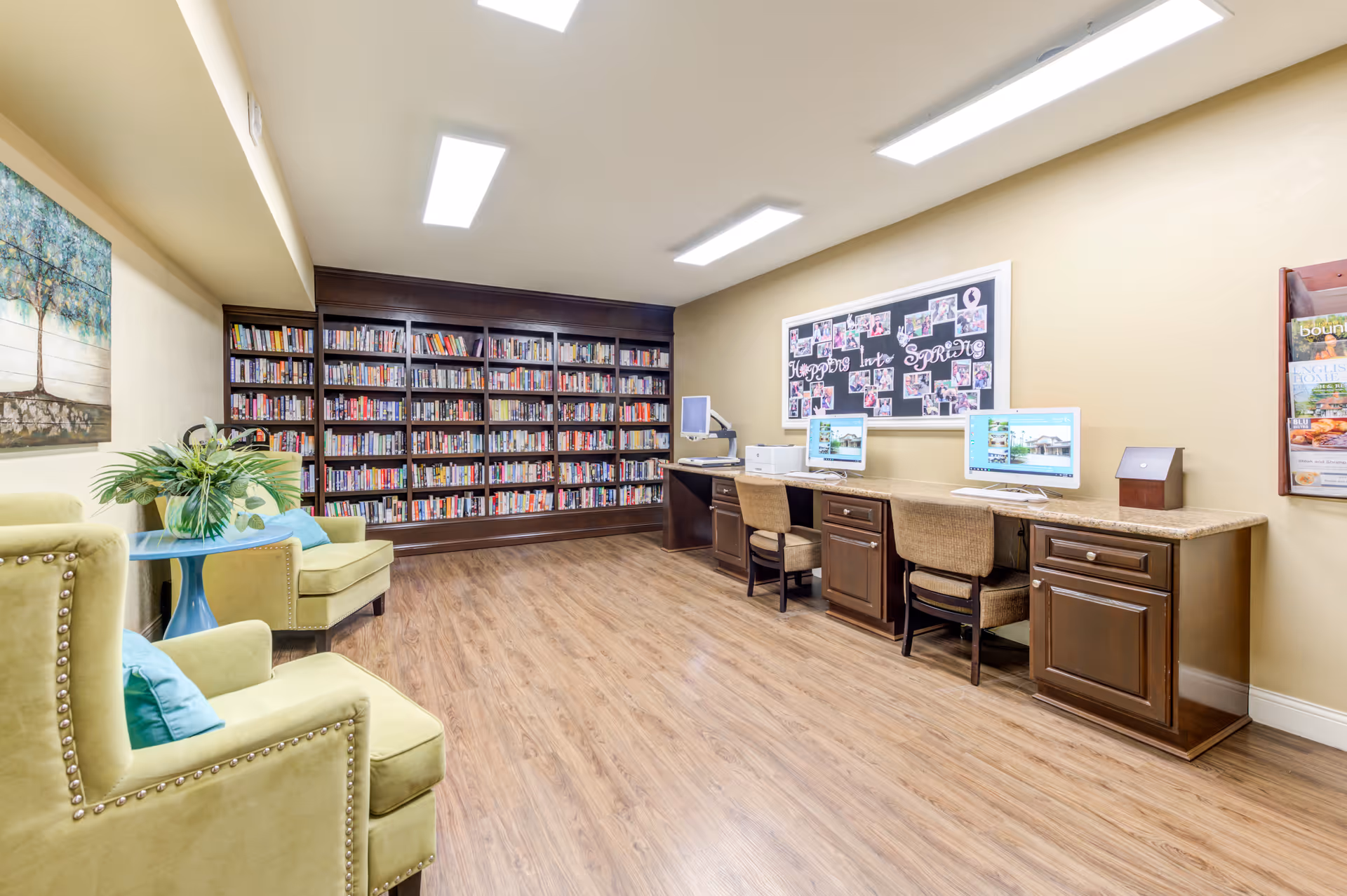 A well-lit room with wooden flooring featuring a large bookshelf filled with books along one wall. There are two green armchairs with blue cushions and a small blue table with a plant on it. Opposite the bookshelf, there is a long wooden desk with two computer monitors, chairs, a printer, and a bulletin board on the wall displaying photos and the text 'Happys Into Spring'.