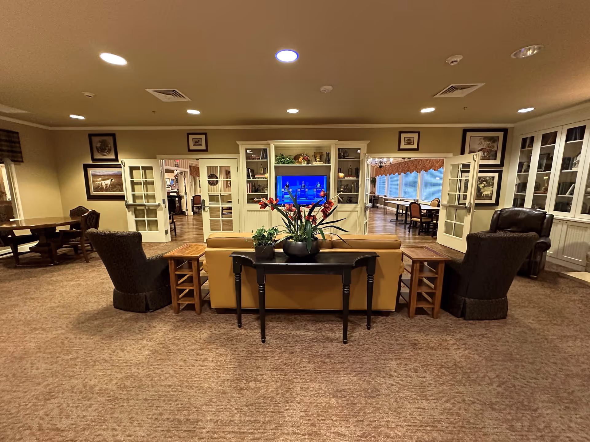 A cozy living room area in a senior living facility with a tan sofa facing a television set in a white entertainment center. Two dark upholstered armchairs flank the sofa, and a black console table with potted plants and flowers is placed behind the sofa. The room has beige walls, carpeted floors, framed pictures on the walls, and recessed ceiling lights. Through open double doors on either side of the entertainment center, a dining area with tables and chairs is visible.