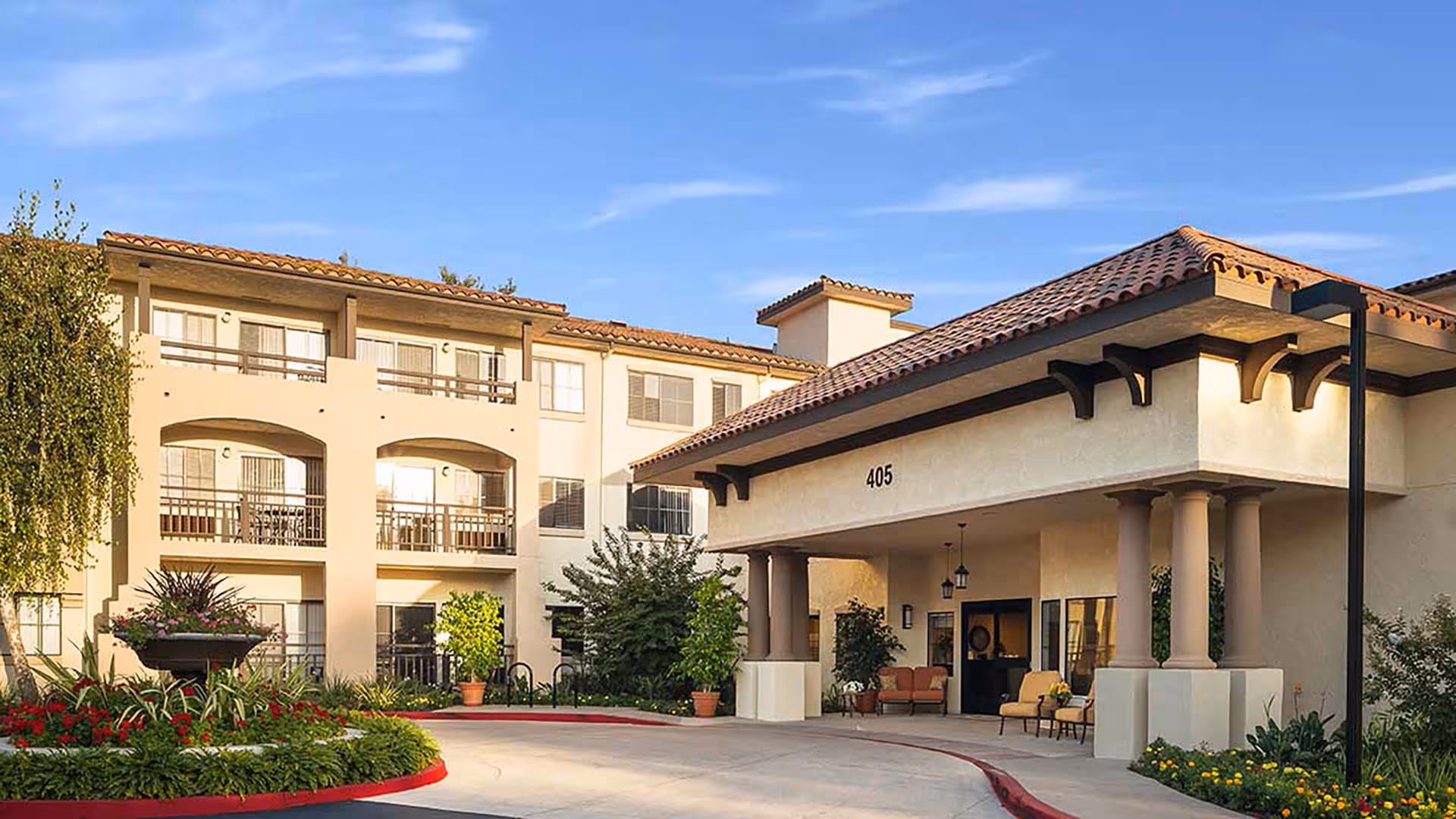 Exterior view of Atria Hillcrest senior living facility showing a three-story building with balconies and a covered entrance area with columns, surrounded by landscaped greenery and a clear blue sky.