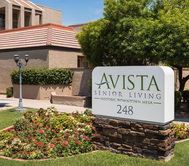 Outdoor view of Avista Senior Living Downtown Mesa sign with the address number 248, surrounded by a landscaped garden with flowers and greenery, and a building with beige walls and a red-tiled roof in the background.