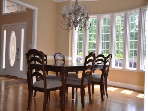 Sunlit dining room with a wooden table and six chairs under a chandelier beside large bay windows and a front door.